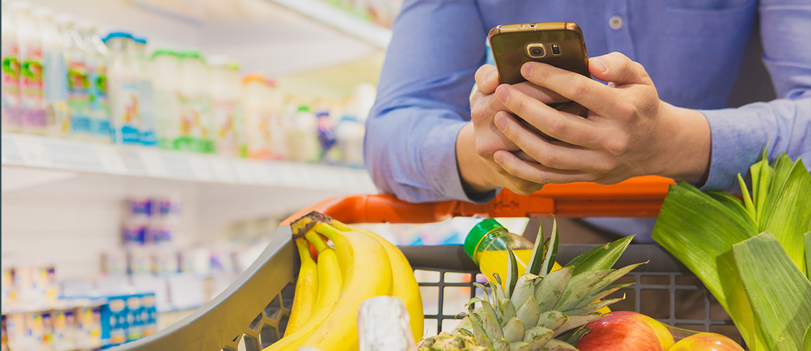 Homem segurando um celular, está no supermercado com um carrinho de compras com frutas, legumes e alimentos. Ao fundo vemos uma geladeira com frios.